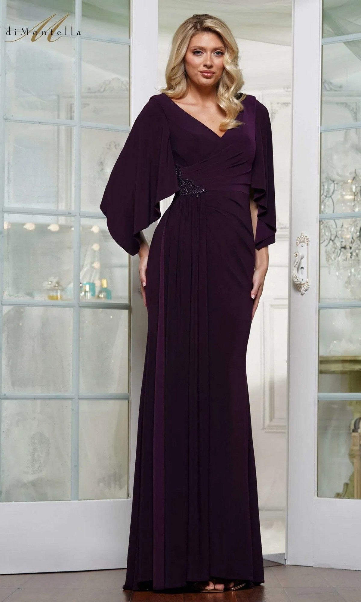 A woman stands indoors wearing the Rina Di Montella RD3238 long formal dress in aubergine, featuring a floor-length silhouette, wrap-style pleated bodice, V-neckline, and draped sleeves.